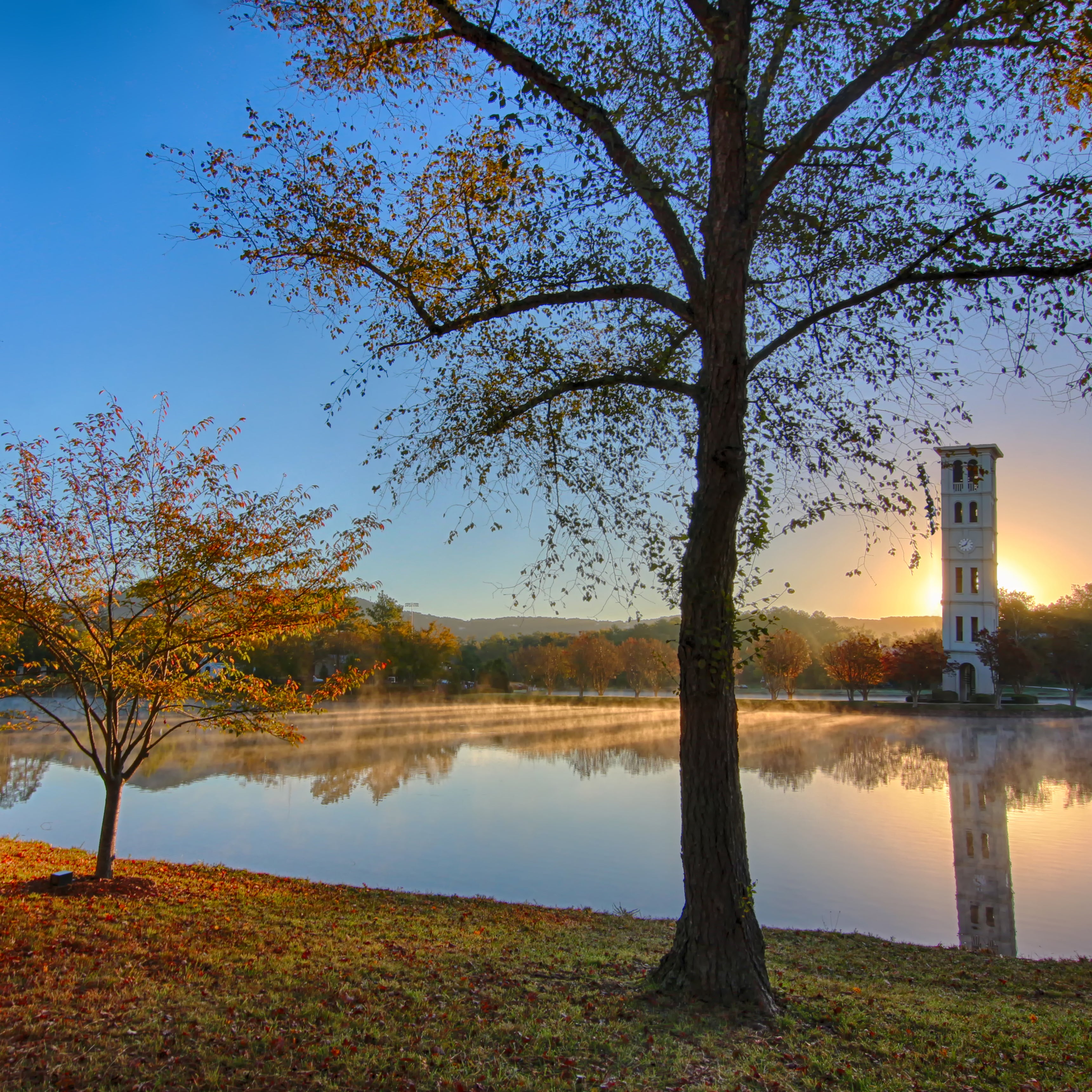 Autumn leaves on the ground by a lake. The sun is rising behind a bell tower.