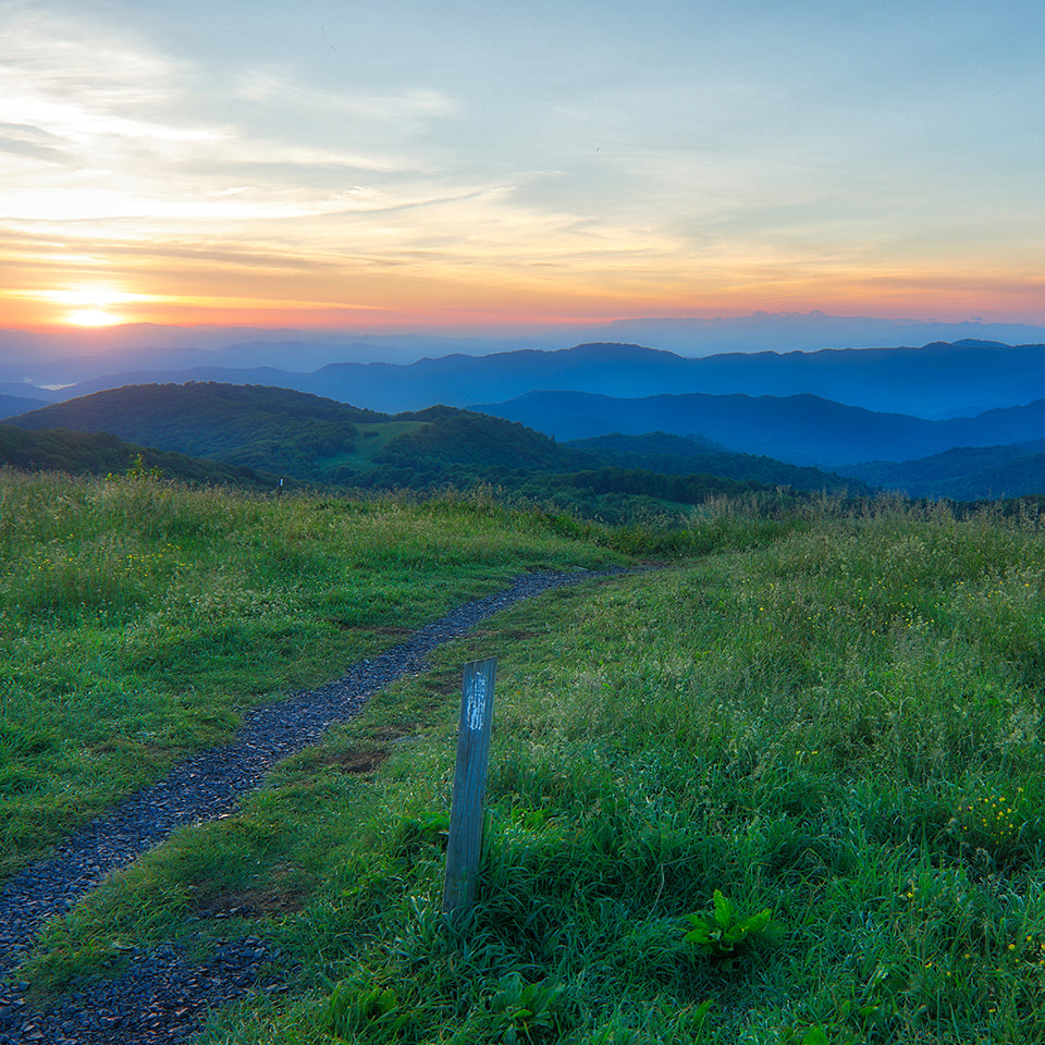 The appalachian trail flows over the mountain with a trail marker at the side.  Gradient colors of sunrise in the distance.