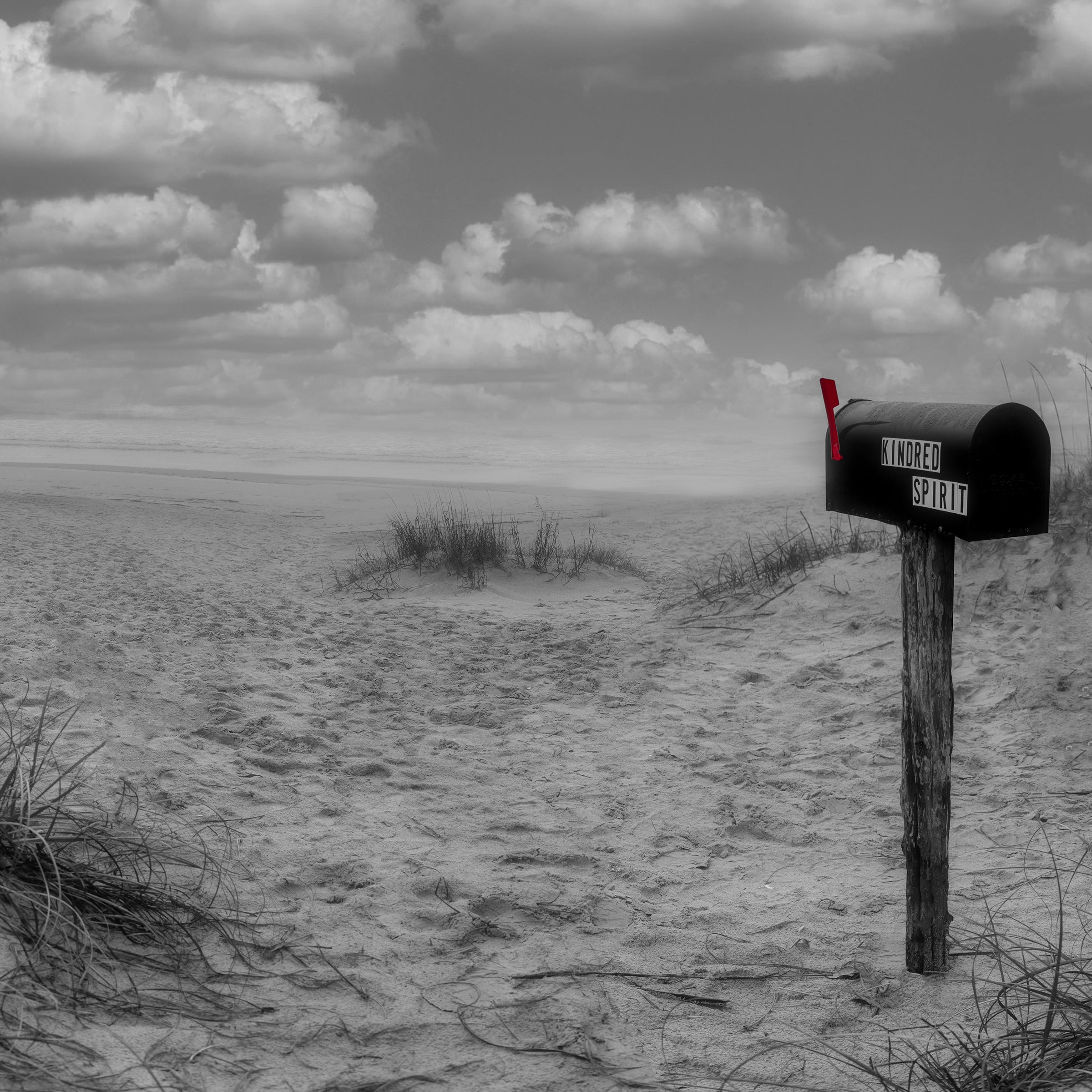 A mailbox on the beach with a label on the side that reads "Kindred Spirit".  Clouds in the sky and a beach background.  The mailbox is not associated with a house but contains journals written by beach goers
